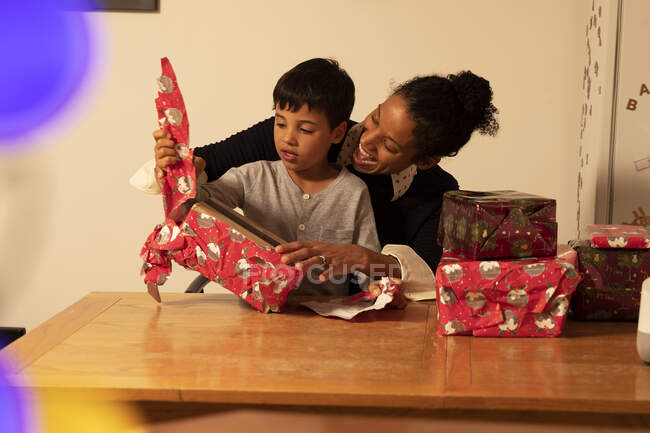 Cheerful mother with son unwrapping Christmas presents on table at home — Stock Photo