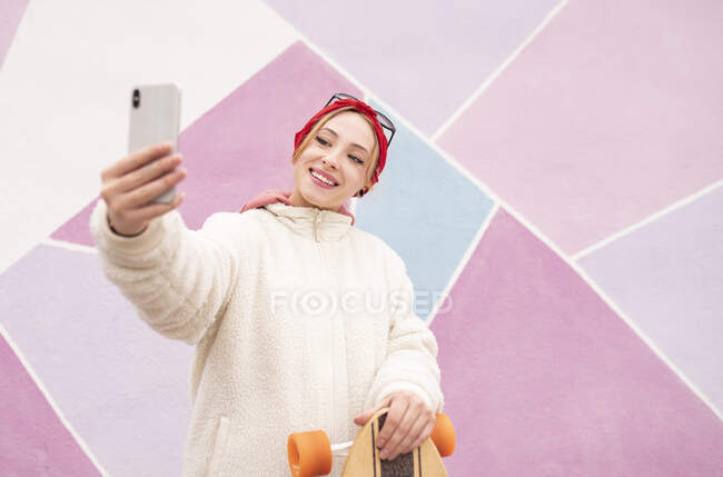 Молодая женщина со скейтбордом делает селфи, стоя напротив разноцветной стены — стоковое фото