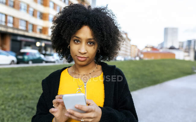 Mujer joven con cabello afro usando teléfono móvil mientras está de pie en la ciudad - foto de stock