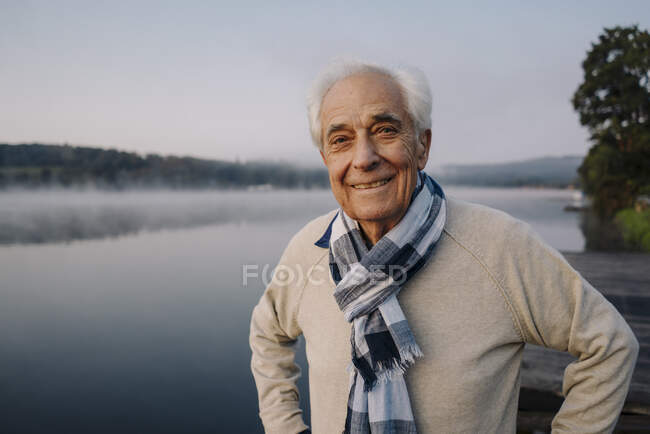 Uomo sorridente indossa sciarpa fissando mentre in piedi contro il lago — Foto stock