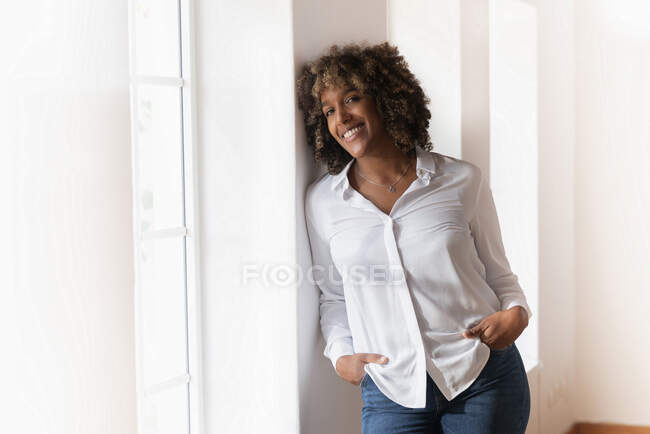 Mid adult woman with hands in pockets leaning on wall while standing by window at home — Stock Photo