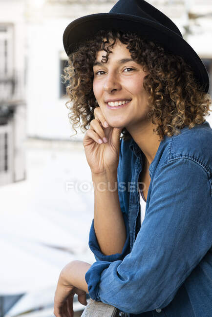 Mujer joven sonriente con sombrero apoyado en pasamanos de balcón - foto de stock