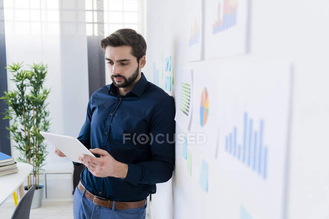 Молодий бізнесмен користується цифровою табличкою, спираючись на стіну в офісі. — стокове фото