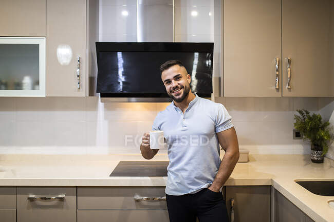 Schöner junger Mann mit Händen in den Taschen, die Kaffeetasse in der Küche haltend — Stockfoto