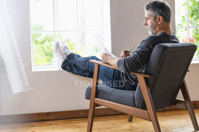 Бизнесмен пишет на бумаге, сидя со скрещенными ногами на кресле дома — стоковое фото