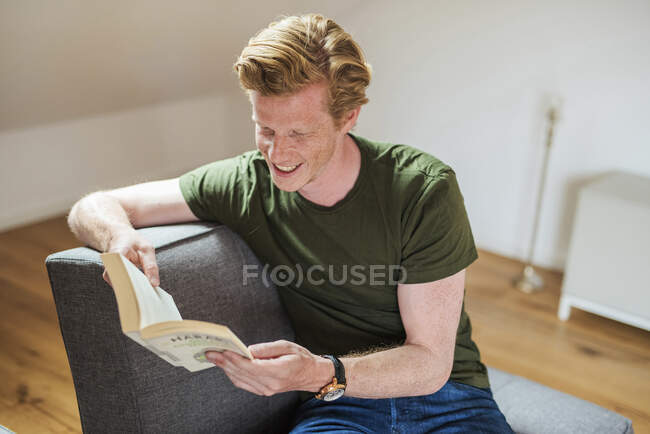 Hombre sonriente leyendo libro mientras está sentado en el sofá en la sala de estar - foto de stock
