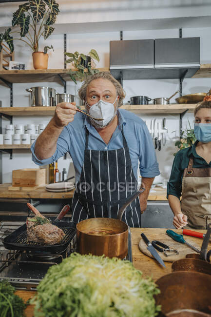 Chef maduro usando máscara facial cozinhar alimentos enquanto está com colega na cozinha — Fotografia de Stock