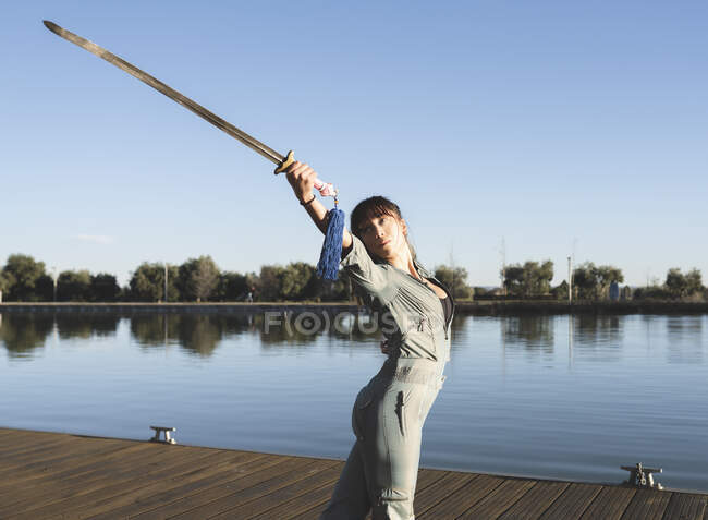 Молода жінка займається бойовими мистецтвами з мечем у парку біля озера в сонячний день. — стокове фото