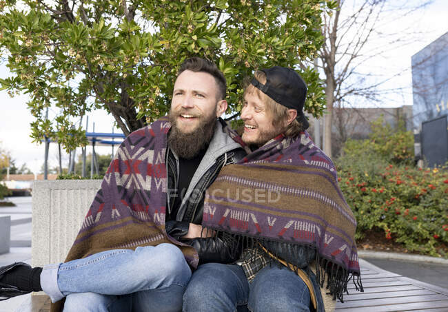 Cheerful gay boyfriends wrapped in shawl sitting on bench — Stock Photo