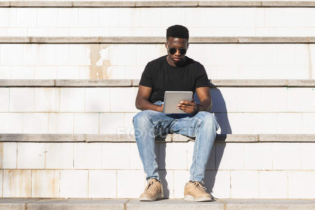 Hombre joven usando tableta digital mientras está sentado en los pasos durante el día soleado - foto de stock