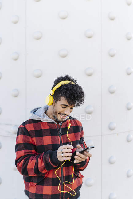 Молодой человек улыбается, используя мобильный телефон, стоящий у стены — стоковое фото