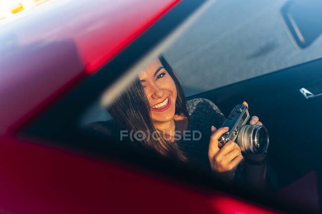 Souriant jeune femme avec caméra vintage à l'intérieur de la voiture vu à travers le pare-brise le jour ensoleillé — Photo de stock