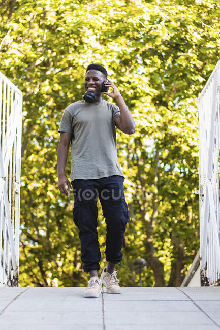 Uomo sorridente che ascolta musica attraverso le cuffie mentre cammina sul ponte pedonale — Foto stock