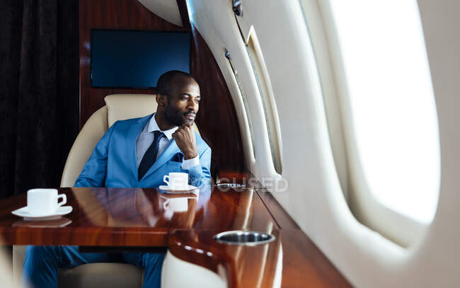 Joven hombre de negocios con la mano en la barbilla mirando a través de la ventana mientras está sentado en jet privado - foto de stock
