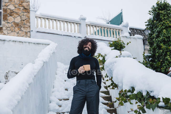 Hombre barbudo sonriente con taza de café de pie en los escalones cubiertos de nieve - foto de stock
