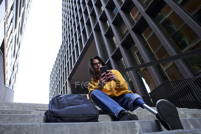 Jeune homme avec un membre artificiel en utilisant un téléphone portable tout en étant assis sur des marches contre le gratte-ciel — Photo de stock