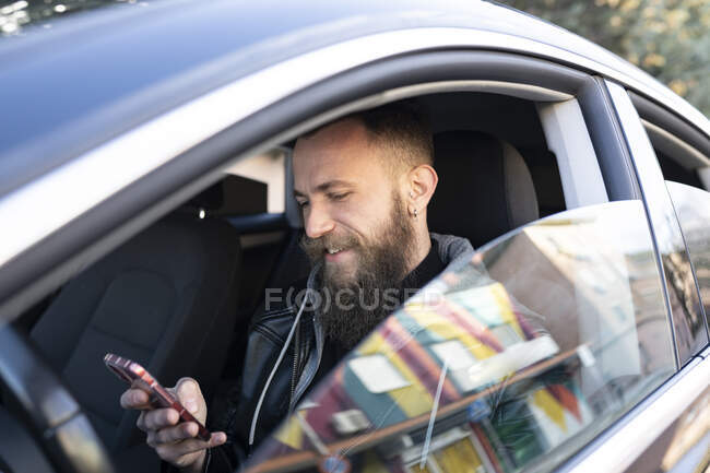 Bärtiger Mann benutzt Handy, während er im Auto sitzt und durch Fenster gesehen wird — Stockfoto
