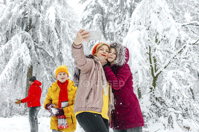 Madre e figlia scattare selfie attraverso il telefono cellulare mentre in piedi con la famiglia in background a foresta — Foto stock