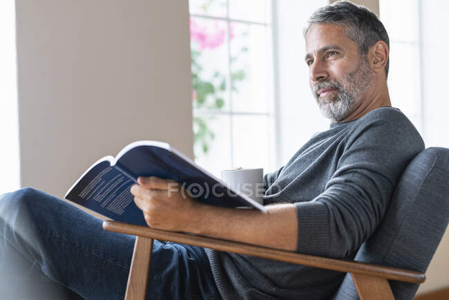 Зрілий бізнесмен з чашкою кави і книгою, дивлячись далеко, сидячи вдома — стокове фото