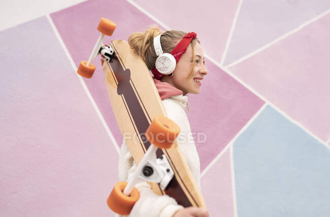 Femme souriante avec écouteurs portant skateboard sur les épaules tout en se tenant contre un mur coloré — Photo de stock