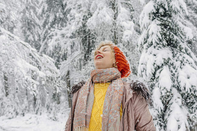 Улыбающаяся женщина наслаждается снегопадом, стоя в лесу — стоковое фото