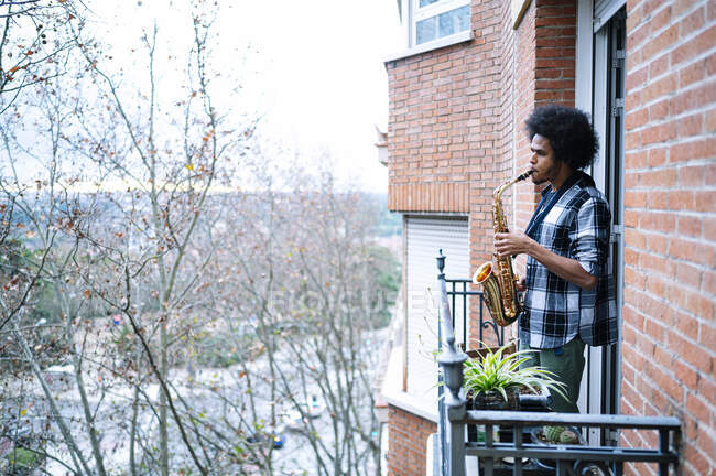 Joven músico tocando el saxofón en el balcón de la casa - foto de stock