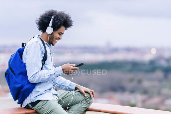 Sonriente joven escuchando música y usando un teléfono inteligente en la terraza contra el cielo - foto de stock