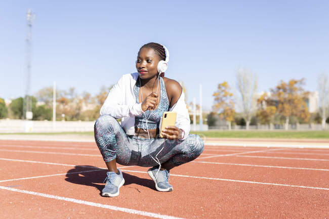Sportiva donna con telefono cellulare che ascolta musica mentre si accovaccia sulla pista sportiva durante la giornata di sole — Foto stock