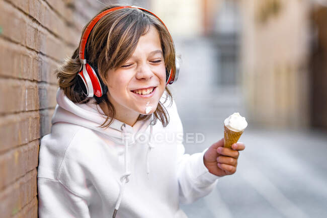 Ragazzo sorridente con le cuffie che tiene il gelato appoggiato alla parete — Foto stock