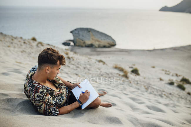 Junger Mann sitzt auf einer Sanddüne während er ein Buch zeichnet in Almeria, Wüste Tabernas, Spanien — Stockfoto