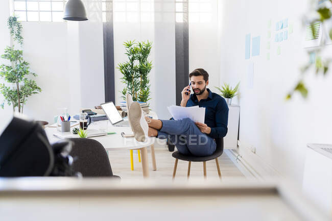 Joven hombre de negocios discutiendo en el teléfono inteligente mientras está sentado con los pies en el escritorio en la oficina - foto de stock