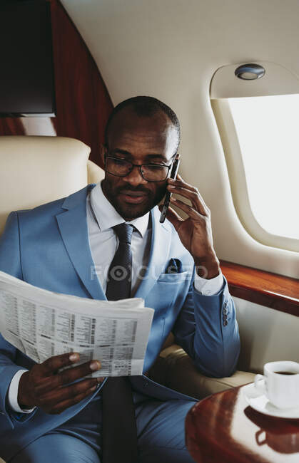 Young businessman reading newspaper while talking on smart phone in airplane — Stock Photo