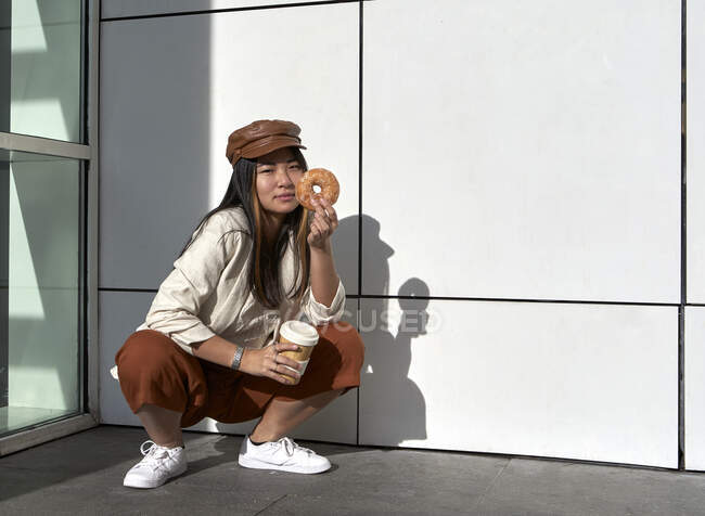 Junge Frau kauert mit Mehrwegbecher und Donut gegen Wand — Stockfoto