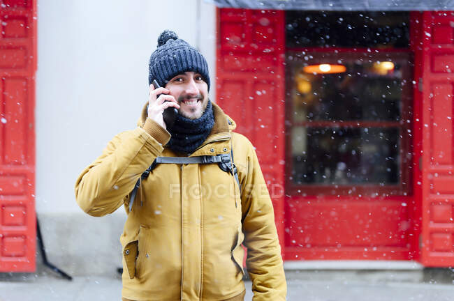 Homme souriant parlant sur un téléphone portable tout en se tenant contre le magasin pendant l'hiver — Photo de stock