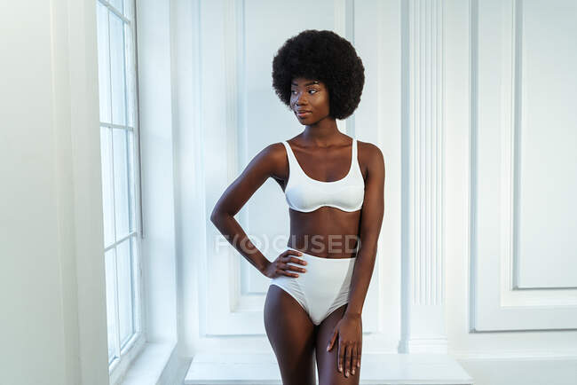 Afro jovem modelo de moda com a mão na anca em lingerie branca olhando para longe contra a parede — Fotografia de Stock