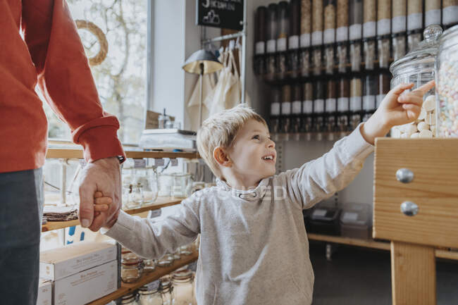 Ragazzo allegro che tiene per mano il padre mentre indica il barattolo di caramelle in negozio — Foto stock