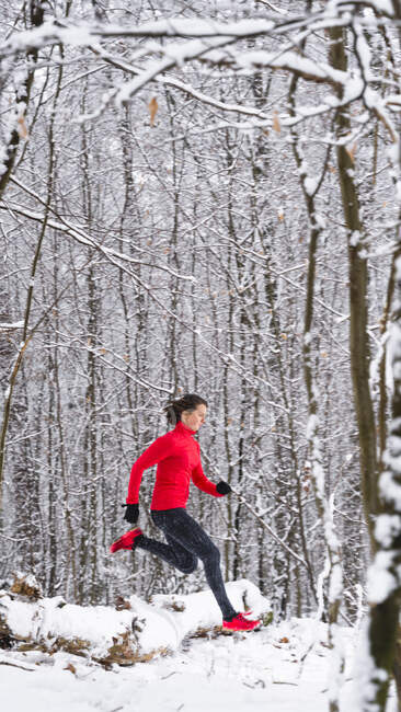 Sportif féminin courant sur un arbre tombé couvert de neige pendant l'hiver — Photo de stock