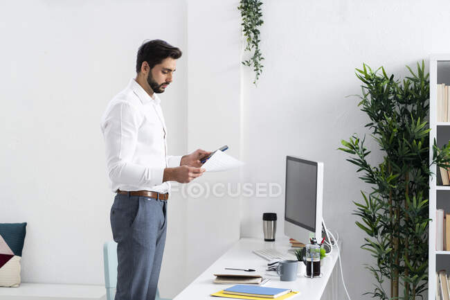 Молодой бизнесмен с бумагой с помощью смартфона на рабочем месте — стоковое фото