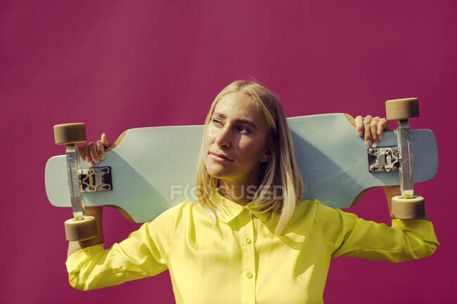 Молода жінка з скейтбордом проти фіолетової стіни в сонячний день. — стокове фото