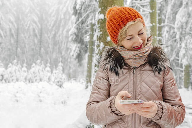 Smiling woman wearing knit hat holding mobile phone while standing in forest during winter — Stock Photo