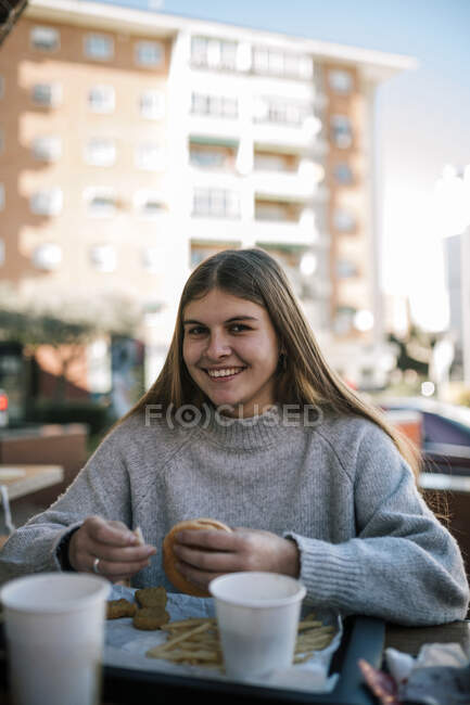 Улыбающаяся девочка-подросток ест бургер, сидя в кафе на тротуаре — стоковое фото