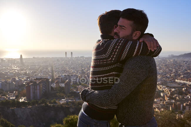 Namorados gays abraçando enquanto estão contra a paisagem urbana durante o nascer do sol, Bunkers del Carmel, Barcelona, Espanha — Fotografia de Stock