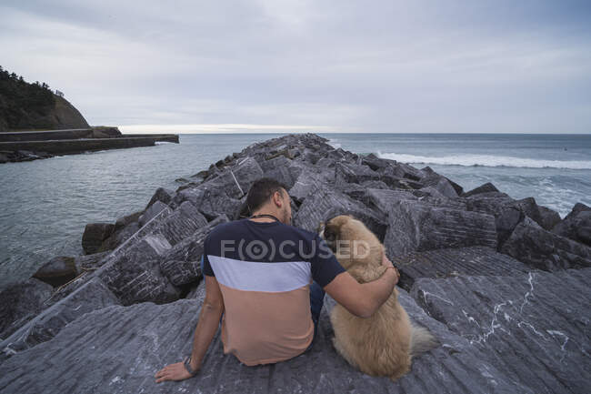 Людина з рукою навколо собаки, сидячи на скелі проти неба. — стокове фото