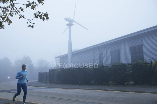 Uomo che corre lungo la nebbiosa strada con turbina eolica sullo sfondo — Foto stock