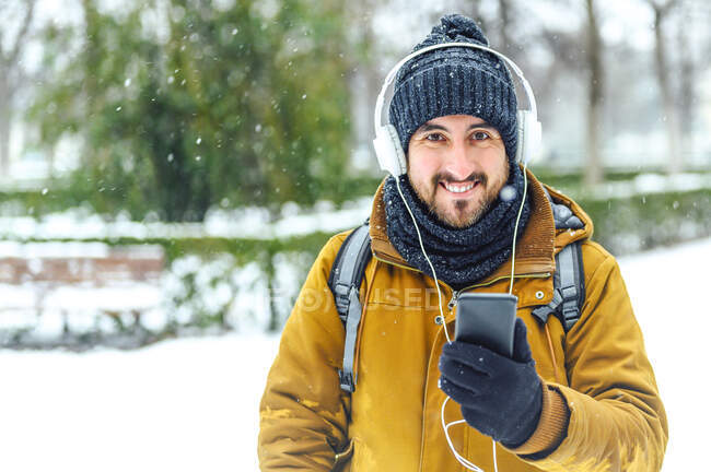 Улыбающийся мужчина держит мобильный телефон, слушая музыку через наушники зимой — стоковое фото