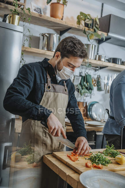 Mid adult chef wearing protective face mask cutting vegetable while standing with colleague in kitchen — Stock Photo