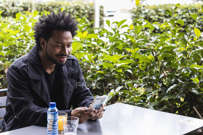 Sonriendo hombre adulto medio utilizando el teléfono inteligente mientras está sentado junto a las plantas en la acera cafetería - foto de stock