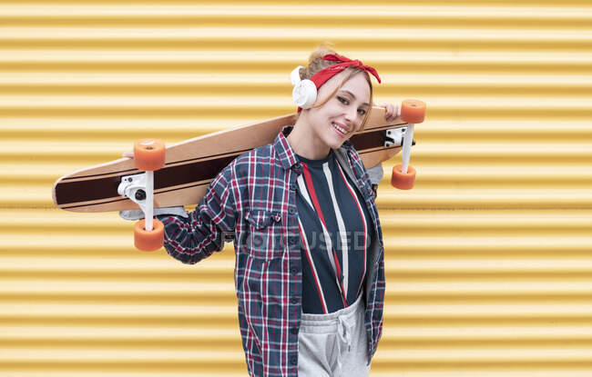 Junge Frau lächelt, während sie Skateboard auf Schulter gegen gelbe Wand trägt — Stockfoto