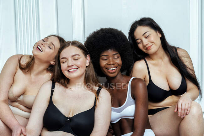 Щаслива багатонаціональна група моделей в білизні з закритими очима сидять біля білої стіни. — стокове фото