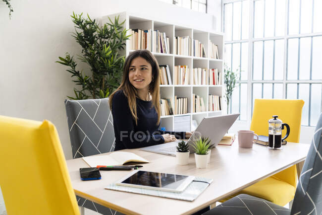 Mujer freelancer sonriente con portátil mirando hacia otro lado mientras está sentada en la sala de estar - foto de stock